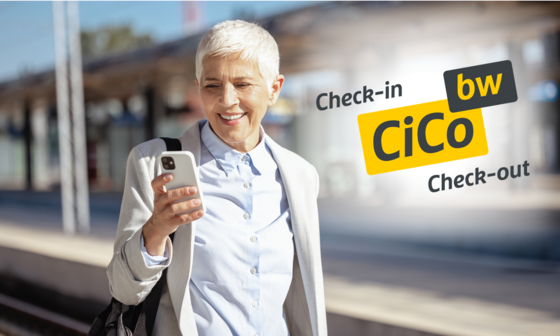 Frau mittleren Alters steht bei schönem Wetter am Bahnsteig und schaut gut gelaunt auf Ihr Handy